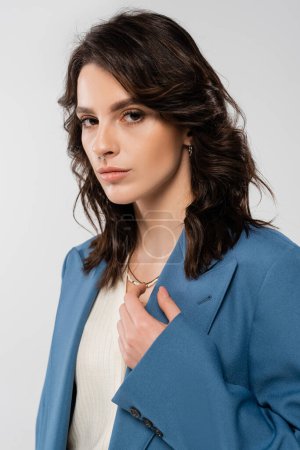 Porträt einer jungen brünetten Frau in blauer, stylischer Jacke, die isoliert auf grau in die Kamera blickt