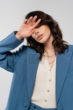 Téléchargez les photos : Femme tendance en veste bleue et colliers couvrant le visage avec main isolée sur gris - en image libre de droit