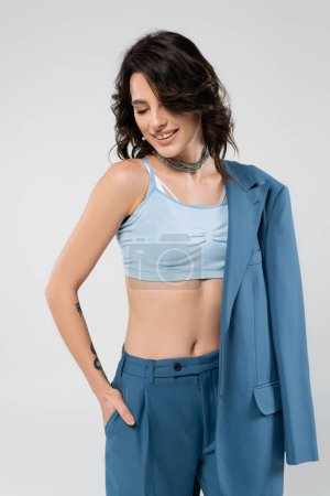 smiling woman in blue crop top standing with hand in pocket of trousers isolated on grey