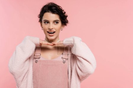 fröhliche Frau im Trägerkleid posiert mit den Händen unterm Kinn und schaut isoliert auf rosa