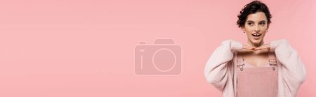 Photo for Excited brunette woman holding hands under chin while posing isolated on pink, banner - Royalty Free Image