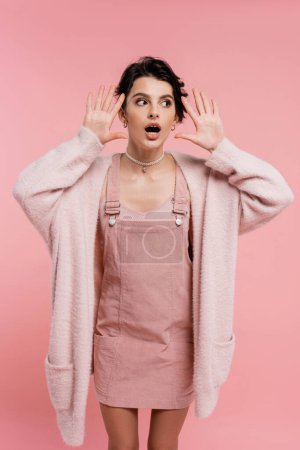 schockierte Frau in Trägerkleid und warmer Strickjacke steht mit offenem Mund und zeigt Wow-Geste auf rosa