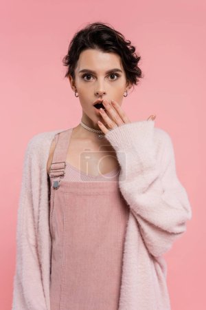 schockierte Frau in Trägerkleid und warmer Strickjacke, die offenen Mund bedeckt, Hand isoliert auf rosa