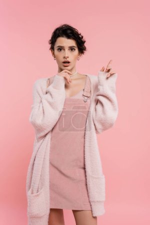 erstaunt Frau in kuscheliger Strickjacke berührt Kinn und zeigt mit dem Finger isoliert auf rosa