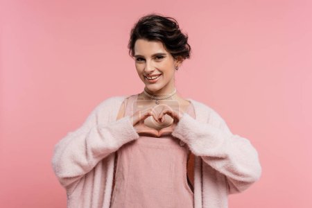 glückliche Frau in warmer und flauschiger Strickjacke mit Herzzeichen und isolierten Händen auf rosa