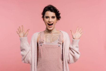 erstaunt und fröhlich Frau in Trägerkleid und Strickjacke zeigt Wow-Geste isoliert auf rosa
