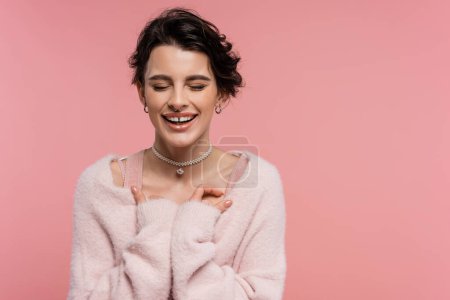 fröhliche Frau in flauschiger Strickjacke und Perlenkette lachend mit geschlossenen Augen isoliert auf rosa