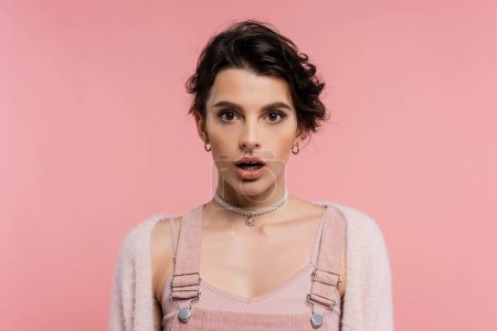 shocked brunette woman with open mouth looking at camera isolated on pink