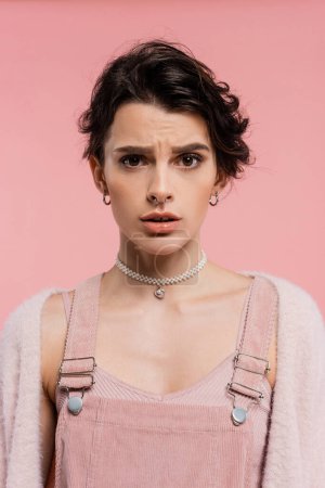 Besorgte Frau in Trägerkleid und Perlenkette blickt vereinzelt in die Kamera auf rosa