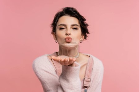 Photo for Pretty brunette woman in cozy cardigan sending air kiss and looking at camera isolated on pink - Royalty Free Image