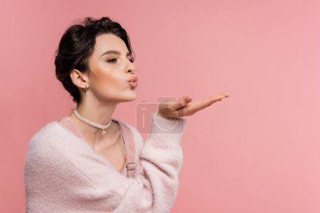 mujer de moda en mullido cárdigan y collar de perlas enviando beso de aire mientras mira hacia otro lado aislado en rosa
