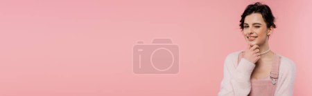 femme délicate et souriante touchant menton et clin d'oeil à la caméra isolée sur rose, bannière