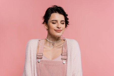 pleased woman in strap dress and cardigan smiling with closed eyes isolated on pink