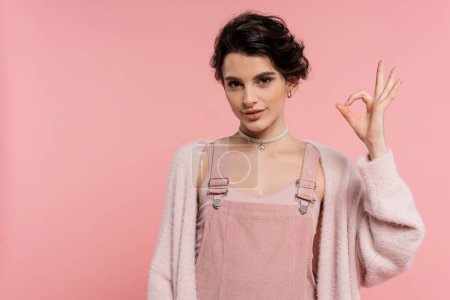 smiling brunette woman in strap dress and fluffy cardigan showing okay sign isolated on pink