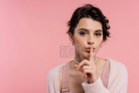 junge brünette Frau zeigt Schweigezeichen und blickt isoliert in die Kamera auf rosa