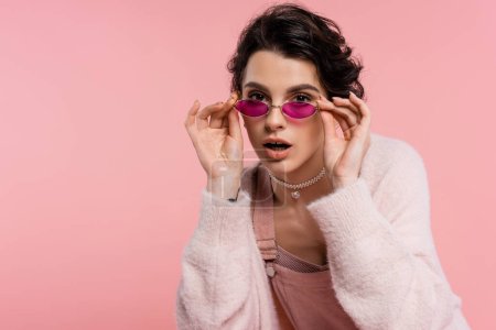 morena mujer en collar de perlas tocando gafas de sol de moda mientras mira la cámara aislada en rosa