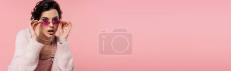 Photo for Young woman adjusting trendy sunglasses and looking away isolated on pink, banner - Royalty Free Image