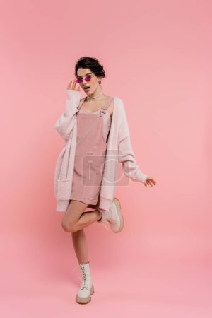 longitud completa de la mujer sorprendida en gafas de sol y chaqueta esponjosa posando en rosa 