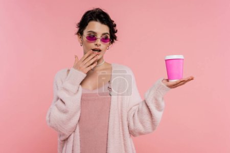 mujer reflexiva en gafas de sol con estilo cogido de la mano cerca de la boca mientras mira la bebida para llevar aislado en rosa