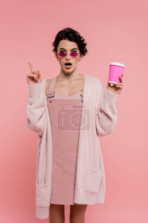 staunte Frau in warmer Strickjacke und Sonnenbrille mit Pappbecher und Zeigefinger auf rosa