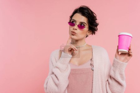 nachdenkliche Frau in trendiger Sonnenbrille mit Imbissgetränk und isoliert auf rosa