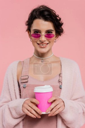 fröhliche Frau in stylischer Sonnenbrille und warmer Strickjacke mit Kaffee to go auf rosa