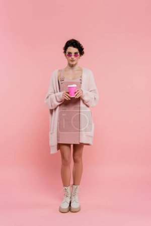 pleine longueur de femme en cardigan chaud et des lunettes de soleil à la mode tenant du café pour aller sur rose 