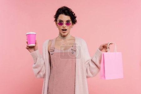 staunende Frau in Sonnenbrille und warmer Strickjacke mit Einkaufstasche und Pappbecher auf rosa