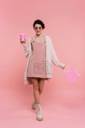 full length of fashionable woman walking with paper cup and shopping bag on pink 