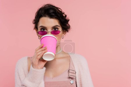 Téléchargez les photos : Brunette femme à la mode lunettes de soleil boire du café de tasse en papier isolé sur rose - en image libre de droit