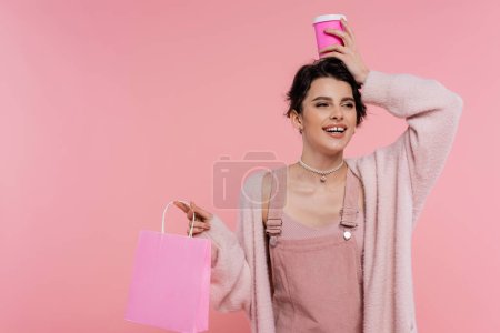 fröhliche und stilvolle Frau mit Einkaufstasche, die Pappbecher über dem Kopf isoliert auf rosa hält