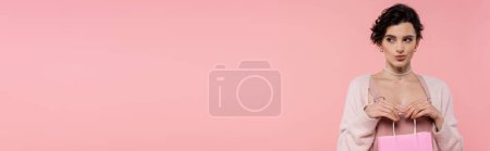 Téléchargez les photos : Femme brune délicate tenant sac à provisions et regardant loin isolé sur rose, bannière - en image libre de droit