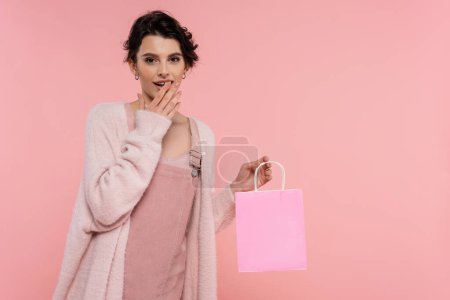 Téléchargez les photos : Femme étonnée dans un cardigan chaud tenant un sac à provisions et couvrant la bouche ouverte avec la main isolée sur rose - en image libre de droit