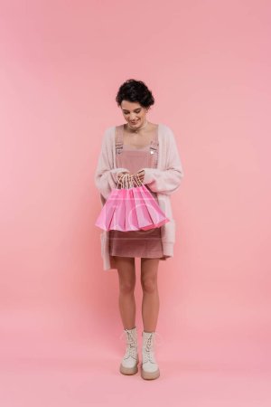 volle Länge der zufriedenen Frau in warmer Strickjacke und Stiefeln mit Blick auf Einkaufstaschen auf rosa Hintergrund
