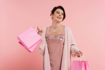 Téléchargez les photos : Femme excitée en robe de sangle et confortable cardigan tenant des sacs à provisions et regardant loin isolé sur rose - en image libre de droit