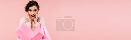 Photo for Amazed brunette woman holding shopping bags near face and looking at camera isolated on pink, banner - Royalty Free Image
