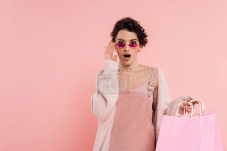 Photo for Astonished woman with shopping bags touching trendy sunglasses and looking at camera isolated on pink - Royalty Free Image