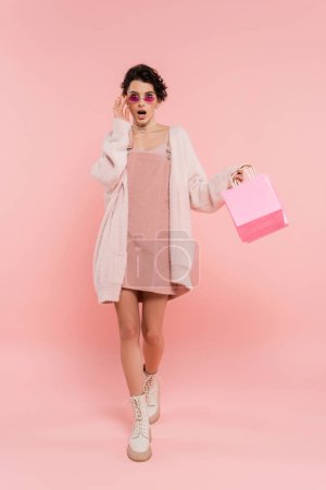fashionable and surprised woman touching sunglasses while walking with shopping bags on pink 