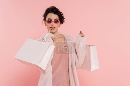 Téléchargez les photos : Femme rêveuse dans des lunettes de soleil à la mode tenant des sacs à provisions et regardant loin isolé sur rose - en image libre de droit