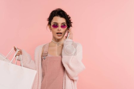 brunette woman in stylish sunglasses holding shopping bags and talking on smartphone isolated on pink