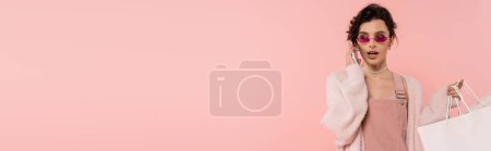 Téléchargez les photos : Femme brune dans un cardigan chaud et des lunettes de soleil tenant des sacs à provisions et parlant sur smartphone isolé sur rose, bannière - en image libre de droit