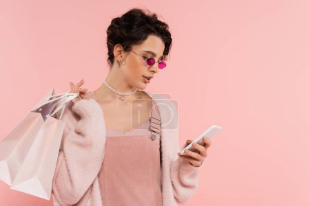 mujer en gafas de sol de moda sosteniendo bolsas de compras y mensajería en el teléfono inteligente aislado en rosa