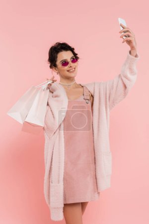 Photo for Happy woman in sunglasses and warm cardigan holding shopping bags and taking selfie on smartphone isolated on pink - Royalty Free Image