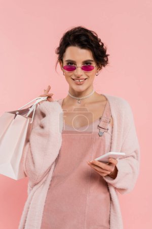 happy and fashionable woman with shopping bags and mobile phone looking at camera isolated on pink