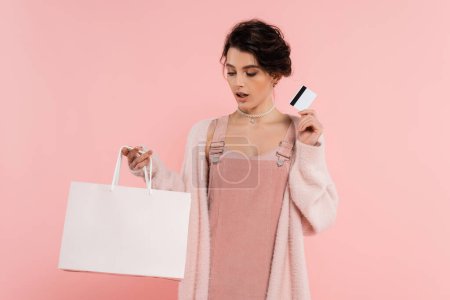 Téléchargez les photos : Femme étonnée en robe de sangle élégante tenant carte de crédit et sac à provisions isolé sur rose - en image libre de droit