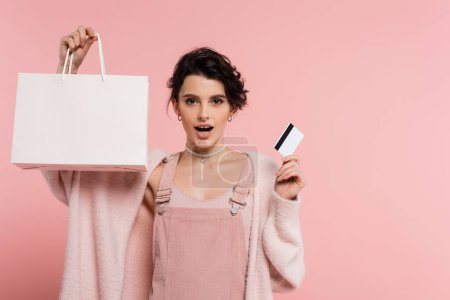 Téléchargez les photos : Femme étonnée avec carte de crédit et sac à provisions en regardant la caméra isolée sur rose - en image libre de droit