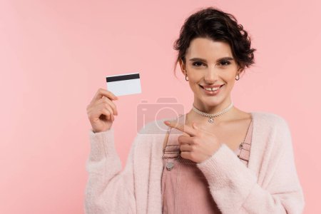 Foto de Alegre morena mujer apuntando a la tarjeta de crédito y sonriendo a la cámara aislado en rosa - Imagen libre de derechos