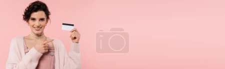 Téléchargez les photos : Joyeuse jeune femme regardant la caméra et pointant vers la carte de crédit isolée sur rose, bannière - en image libre de droit
