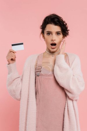 Téléchargez les photos : Femme excitée dans le cardigan chaud tenant la carte de crédit et la main près du visage isolé sur rose - en image libre de droit