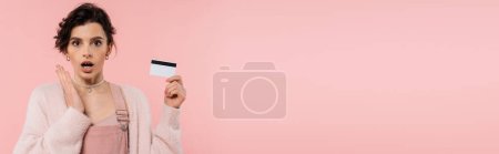 Téléchargez les photos : Femme étonnée dans le cardigan chaud tenant la carte de crédit et regardant la caméra isolée sur rose, bannière - en image libre de droit